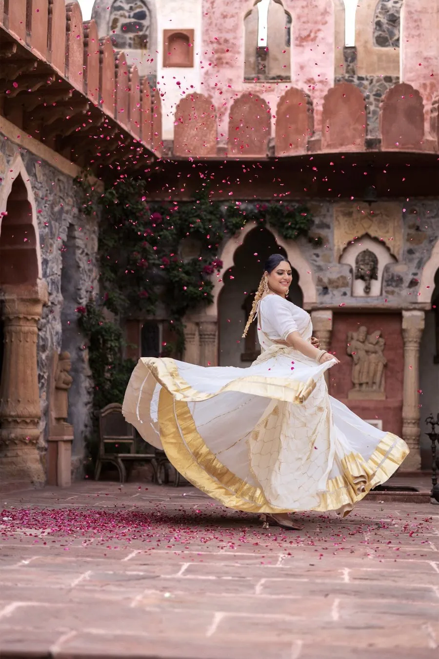 Mahi twirling in a white and gold saree in a palace courtyard, rose petals falling around her